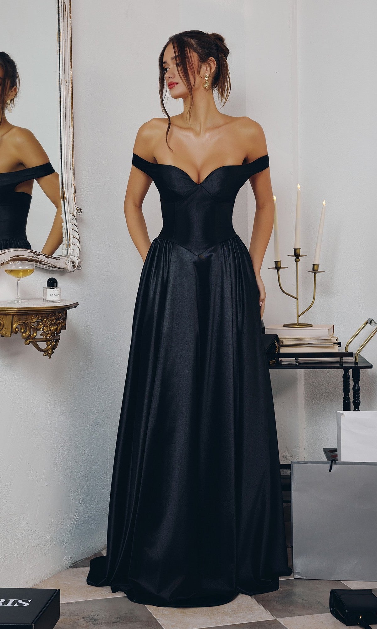 A woman in the Amoris T821 black long prom dress stands indoors near a mirror and a table with candles and boxes, showcasing the timeless elegance of Cinderella Divine.